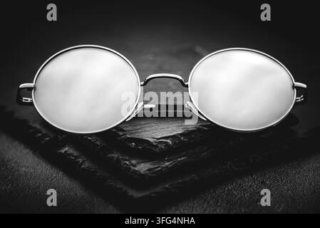High-contrast black and white close-up of round wire-rimmed spectacles resting on a dark slate background, symbolizing vision or intellect. Stock Photo