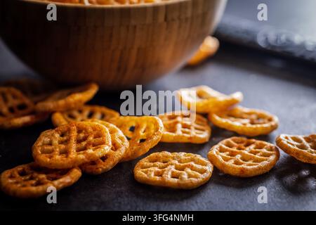 Crispy, seasoned waffle-cut potato chips scattered on a dark tabletop with a wooden bowl in the background, highlighting golden texture and savory cru Stock Photo