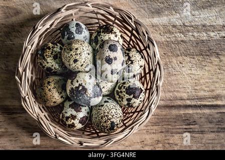 Top view flat lay of speckled quail eggs nestled inside a small woven wicker basket on a rustic wooden background with copy space. Stock Photo