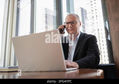 Mature businessman talks on smartphone while using laptop in modern office, representing multitasking, client communication and digital work. Stock Photo