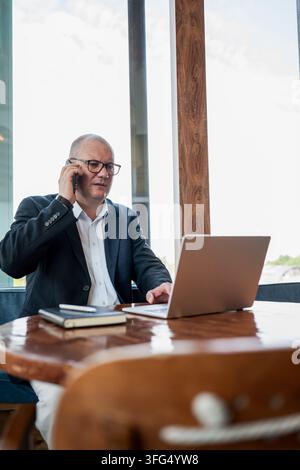 Mature businessman talks on smartphone while working with laptop at office table, with large copy space for business advertising. Vertical format. Stock Photo