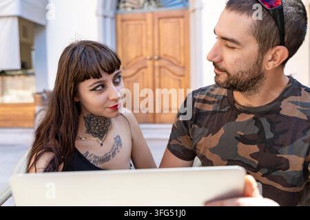 Young couple sitting outdoors, interacting and talking while using a digital tablet at cafe Stock Photo
