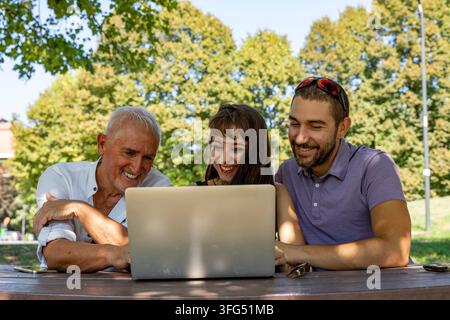 Happy people of different generations using a laptop together at an outdoor table in park Stock Photo