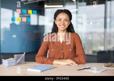 Young businesswoman smiling at camera, sitting at a desk in a contemporary office setting, representing professionalism, success, and a positive workplace environment Stock Photo