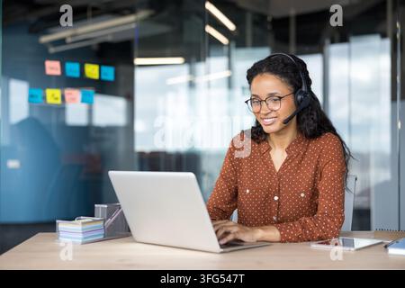 Smiling young woman wearing a headset and glasses working on a laptop in a modern office, providing telemarketing assistance and client communication services Stock Photo