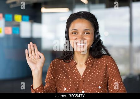 Happy call center agent wearing headset and smiling at camera, making an inviting hand gesture in a modern office, conveying friendly customer service and professional support Stock Photo