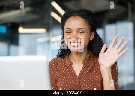 Woman smiling and waving hand during a video conference on a laptop, connecting remotely from an office environment, engaging in communication and a virtual greeting Stock Photo