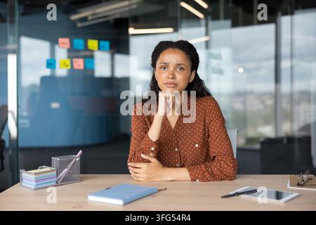 Young professional african-american woman sitting at a desk in a contemporary office, appearing thoughtful and concerned, contemplating a challenging problem while looking directly at the camera Stock Photo