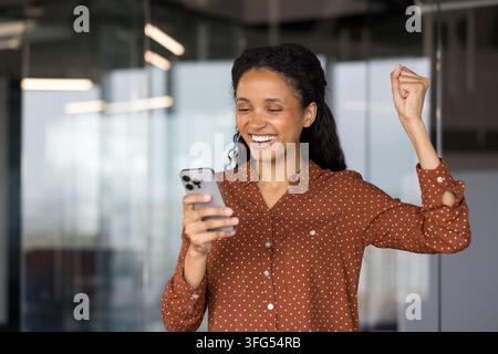 Young woman looking at her mobile phone and celebrating professional success, receiving good news, or achieving a goal, expressing excitement and happiness in an office environment Stock Photo