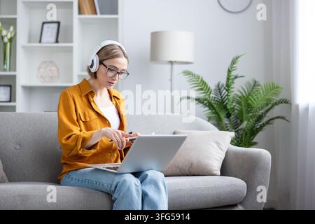 Young woman sitting on a comfortable sofa, wearing headphones and glasses, concentrating on her laptop while working from home or engaging in online education Stock Photo