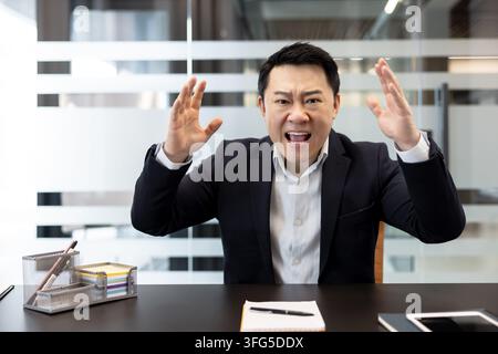 Asian businessman expressing strong frustration and anger, yelling with hands raised, sitting at a desk in a corporate office setting, feeling stressed and overwhelmed Stock Photo