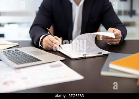 Business professional is signing a document on a clipboard. Holding another paper. With a laptop and financial charts on a dark office desk. Representing agreement. Paperwork. And official procedures Stock Photo