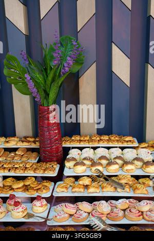A colorful dessert buffet featuring a wide selection of fresh pastries, glazed donuts, mini cakes, and sweet rolls arranged on serving trays. In the b Stock Photo