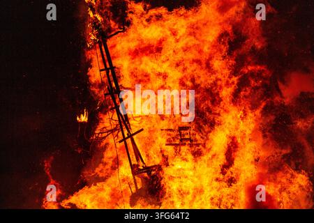 WOODEN EFFIGY ENGULFED IN RAGING FLAMES, BURNING THE WICKER MAN EFFIGY, GREEN MAN FESTIVAL, BRECON, WALES, UK, 2016: A close-up aerial view shows a wooden boat-like structure being consumed by intense orange and yellow flames against a dark night sky. Photo: Rob Watkins. INFO: The Green Man Festival is held annually in the Brecon Beacons in August. It celebrates music, arts, literature, science and family-friendly culture in a beautiful mountain setting. Each year the festival ends with the ritual burning of the wicker man Green Man effigy. Stock Photo