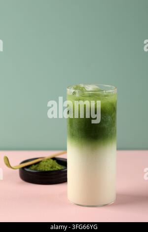 Green matcha latte iced drink in tall glass. Pink surface, mint backdrop, minimalist aesthetic. Fresh ice, creamy layer, vibrant green top. Tea spoon Stock Photo