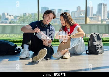 Two young students studying together with notebooks sitting on floor near big window Stock Photo