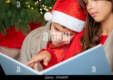 Brother and Sister Reading Christmas Book Together by Festive Tree at Home Stock Photo