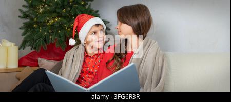 Brother and Sister Reading Christmas Book Together by Festive Tree at Home Stock Photo