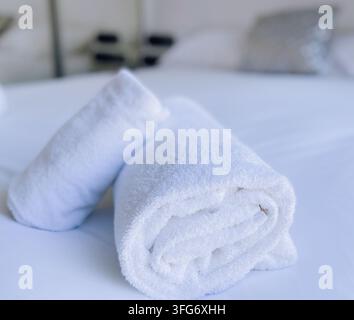 Fresh rolled white bath towels arranged neatly on a clean white bed sheet. Stock Photo