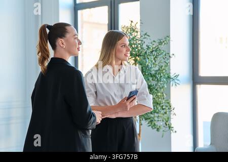 Two business women standing near window looking away talking in modern office Stock Photo