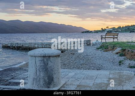 An empty iron bench overlooks a calm sea and distant mountains at sunset, framed by a stone pier, pebbled shore and soft pastel sky — a peaceful coast Stock Photo