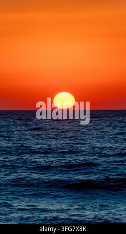 Bright sun setting directly over the sea horizon beneath an intense orange sky, with dark blue ocean waves creating strong warm and cool color contras Stock Photo