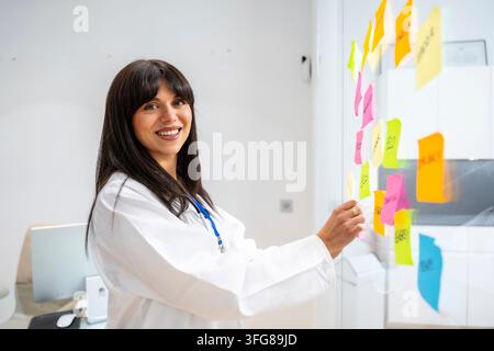 Female doctor in a white coat smiling confidently, organizing and analyzing various healthcare concepts and ideas written on colorful sticky notes app Stock Photo