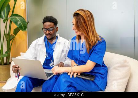 Diverse medical professionals wearing scrubs and lab coat collaborating while reviewing results on a laptop, emphasizing teamwork and communication in Stock Photo