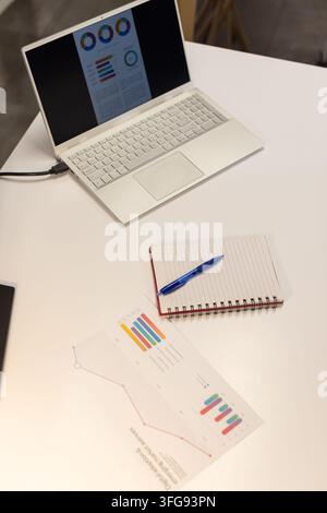 Open laptop showing charts on table, charger trailing, notebook beside printout, backdrop blurring Stock Photo