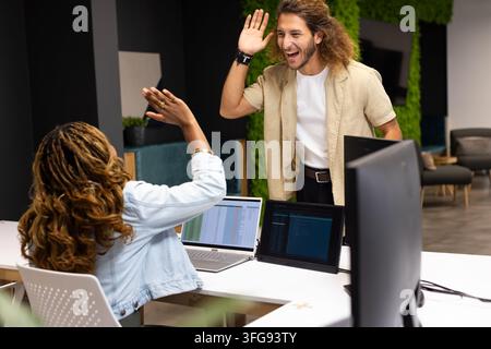 Diverse coworkers high-fiving while working at open-plan office with laptop, denim jacket Stock Photo