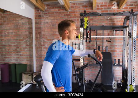 Man standing in home gym by air bike and power rack, wearing blue shirt, white sleeves Stock Photo