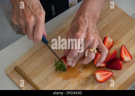 Senior woman slicing strawberries with blue paring knife on wooden board at counter with gold ring Stock Photo