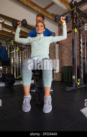 African American woman wearing mint top doing seated dumbbell press at gym with trainer spotting Stock Photo