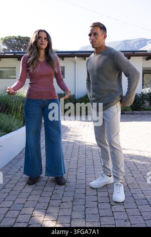 Diverse couple standing on paved courtyard in front of modern house wearing sweater and jeans. Outdoor, residential, two, sunny, minimalist, architect Stock Photo