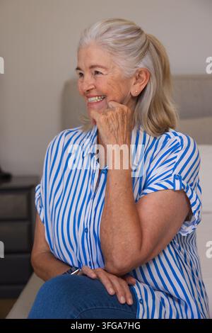 Senior woman sitting on bed in bedroom, smiling and touching chin in striped shirt showing ring Stock Photo
