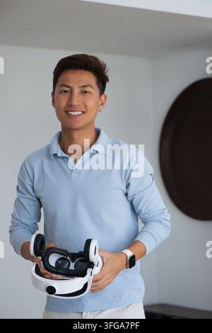 Asian man standing, smiling in studio wearing polo and smartwatch, holding white black VR headset Stock Photo