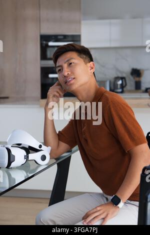 Middle aged Asian man wearing polo sitting at glass table in kitchen touching temple near headset Stock Photo