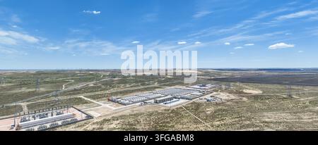 Aerial view of a battery energy storage station and electrical substation in an arid desert region. Vast solar photovoltaic arrays, transmission towers and distant wind turbines spread across the dry landscape under a bright blue sky with scattered clouds. Stock Photo