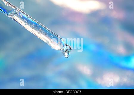 Close-up of a glass dropper releasing a clear serum drop against a blue bokeh background Stock Photo