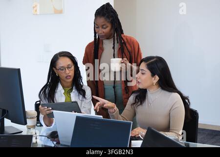 Diverse female coworkers collaborating around glass table in office with laptops, tablet, mug Stock Photo