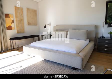 Upholstered platform bed is sitting centered in master bedroom with white duvet, sunlight casting Stock Photo
