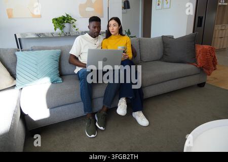 Couple sitting on grey sofa using silver laptop, woman holding ceramic mug, looking in living room Stock Photo