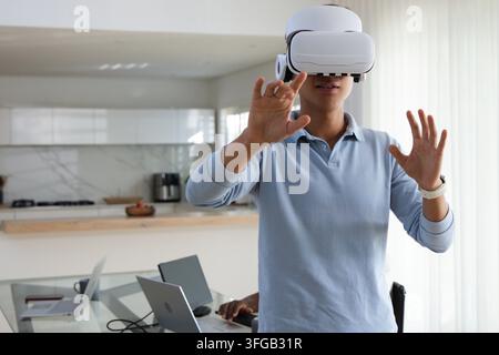 Diverse male coworkers testing white VR headset and typing on laptops at glass table in kitchen Stock Photo