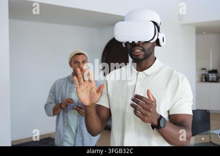 Diverse male coworkers testing VR at home one wearing white headset raising hand Stock Photo
