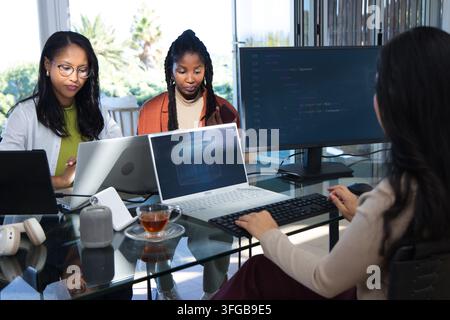 Diverse female colleagues collaborating on code at glass table with three laptops, external monitor Stock Photo
