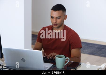 African American man typing on laptop at home glass-top desk with mint-green mug wearing rust shirt Stock Photo