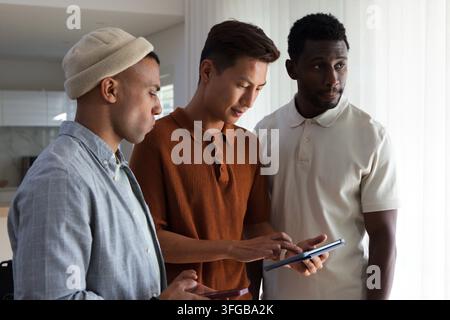 Diverse male coworkers leaning in examining tablet screen in bright open-plan apartment kitchen Stock Photo