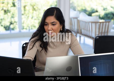 Woman sitting at desk in bright home office wearing beige ribbed top, focusing on multiple laptops Stock Photo