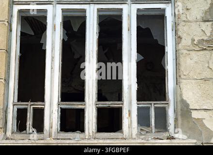 Derelict building facade with weathered white window frames and shattered panes, evoking urban decay, abandonment and neglect in a textured, aged exte Stock Photo