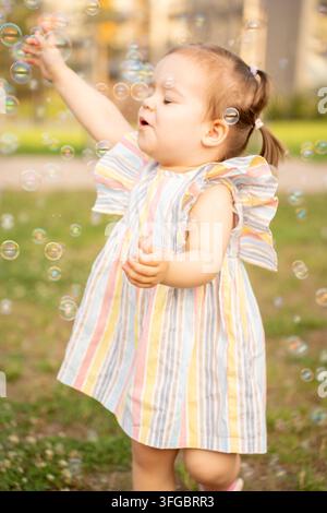 joyful child blowing soap bubbles outdoors in warm summer light, with a softly blurred green garden behind, pastel striped clothing, iridescent sphere Stock Photo